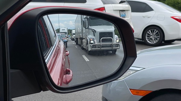 A tractor-trailer in the rear-view mirror of a car stuck in traffic.