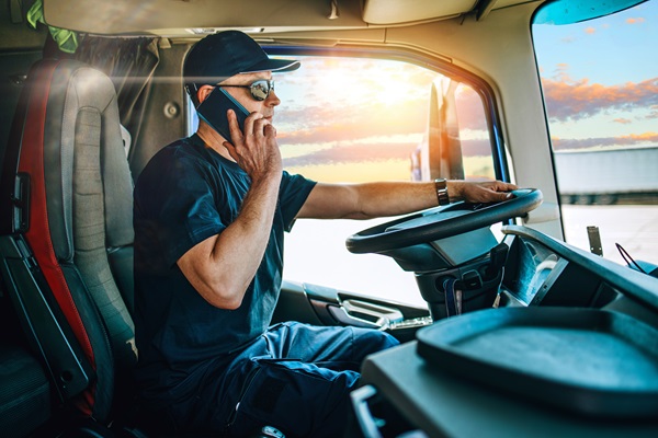 A male truck driver wearing a hat and sunglasses talking on a cell phone held to his ear while driving a commercial vehicle during sunset. (466748050)
